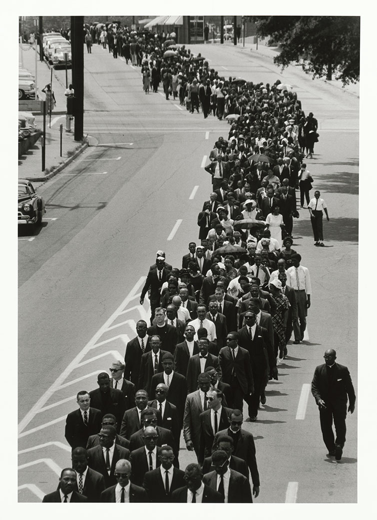 A black-and-white photograph of a crowd of people dressed in suits, or funeral attire, walking in a procession through the streets of Jackson, Mississippi.